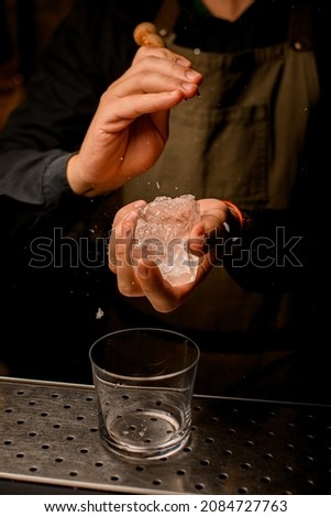 close-up of ice cube in the hands of man. Bartender crushing big ice cube with ice crusher with wooden holder. Empty glass on bar