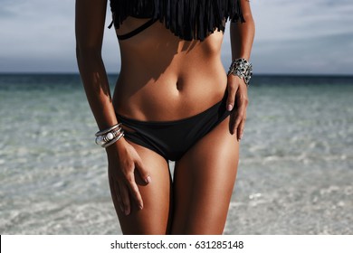Close-up of an ideal fitness body of a luxurious girl on the beach, sexy black bikini, perfect bronze tan skin, Fashionable silver accessories,  low key photo, Radiant skin, Fashion glamor, life style