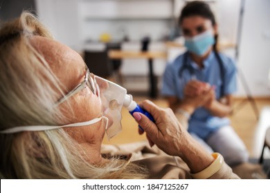 Closeup Of Ill Blond Senior Woman Sitting At Home And Taking Oxygen From Respirator While Nurse With Protective Mask On Sitting Next To Her And Holding Her Hand During Corona Virus.