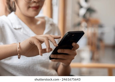 A close-up image of an Asian woman in casual clothes sitting on the sofa in the living room, using her smartphone to check messages or read an online blog. people and wireless technology concepts - Powered by Shutterstock