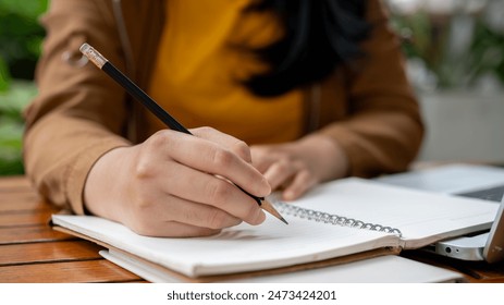 A close-up image of an Asian woman holding a pencil, writing something in her notebook, making list, drawing, taking notes, doing homework, or keeping diary. - Powered by Shutterstock