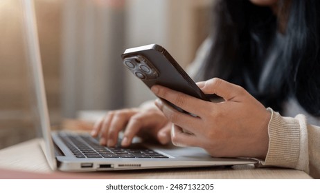 A close-up image of an Asian woman using her smartphone while working on her laptop computer in a coffee shop, working remotely, multitasking. people and wireless technology concepts - Powered by Shutterstock