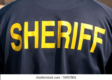 Closeup Image Of A Back Of A Sheriff's Deputy, With Text Sheriff In Yellow Letters