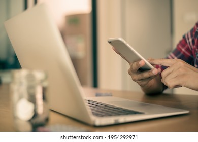 Closeup Image Of A Beautiful Asian Man Holding, Using And Looking At Smart Phone And Checkin Email With Laptop Place On Old Wooden Table At Home.