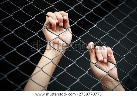 A close-up image capturing hands gripping a metal fence, symbolizing struggle and confinement. The dark background enhances the feeling of desperation and emotion.