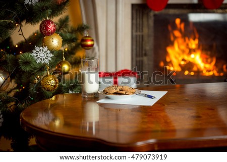 Closeup image of cookies, glass of milk and letter for Santa Claus on table next to burning fireplace and Christmas tree