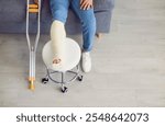 Close-up of the image of crutches and broken leg in a plaster cast. Young man with an injured leg and foot sitting on the couch at home, with his foot resting on a stool. Cropped shot, top view.