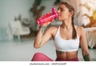 Close-up image of cute woman in sportswear sitting on the floor after exercises or gym and drinking water from the pink sports bottle. Sports trainer or teacher of Pilates - Powered by Shutterstock