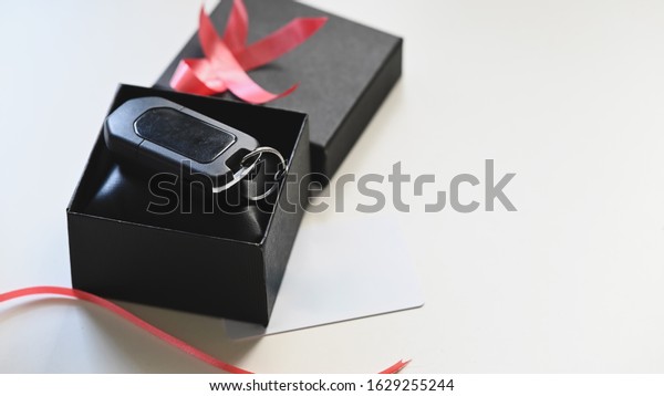 Close-up image\
of the Digital car key putting inside the black gift box with red\
ribbon and wish card on the white desk as background. Surprising\
Valentine\'s Day gift\
concept.