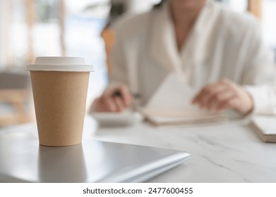 A close-up image of a disposable coffee cup on a white marble table in a coffee shop with a blurred background of a woman working at the table. - Powered by Shutterstock