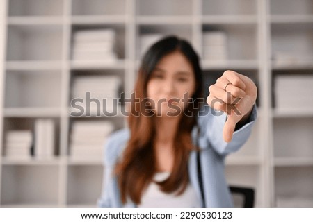 Close-up image of a dissatisfied and upset millennial Asian businesswoman showing thumbs down dislike gesture while sitting at her desk. discontent, disapproval, dissatisfied, displeased