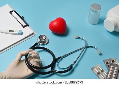 Close-up Image Of Doctor Hand With Glove Holding A Stethoscope Over The Blue Table Surrounded By A Heart Model, Digital Thermometer, Clipboard, Pill Bottle And Capsule Pills.