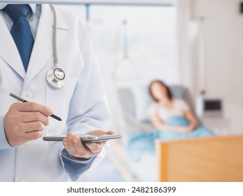 A close-up image of a doctor in a white coat and stethoscope using a digital tablet and stylus. In the blurred background, a patient lies in a hospital bed, suggesting a hospital or clinical setting. - Powered by Shutterstock