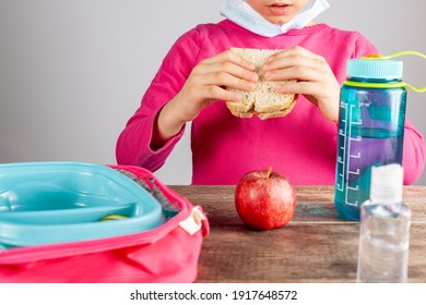 Closeup image with eating lunch at school concept during the phased reopening after COVID-19 pandemic closures. Girl with face mask removed eats sandwich snack with hand sanitizer on the table - Powered by Shutterstock