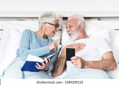 Closeup image of elderly couple lying in bed before sleep. She took a book and he hold smartphone. Both laughing together. Concept of Concept of peaceful married life - Powered by Shutterstock