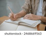 A close-up image of a female college student in comfy clothes holding a pencil, writing something in a notebook at a table indoors. doing homework, keeping diary, making to do list