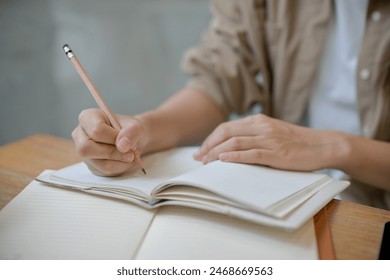 A close-up image of a female college student in comfy clothes holding a pencil, writing something in a notebook at a table indoors. doing homework, keeping diary, making to do list - Powered by Shutterstock