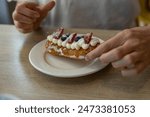 A close-up image of a freshly baked pastry topped with whipped cream and berries, held in the hand of a baker. The pastry is adorned with strawberries and blueberries.