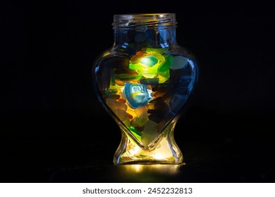 A closeup image of a glass jam jar filled with small pieces of different coloured sea glass. The jar is heart shaped and set against a black background with lights glowing through the glass. - Powered by Shutterstock