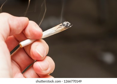 Close-up Image Of A Hand Holding A Lit Marijuana Joint Against Dark Gray Background