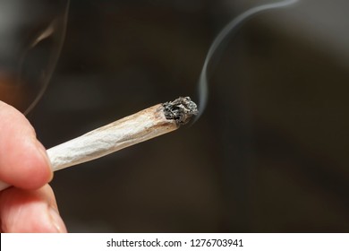 Close-up Image Of A Hand Holding A Lit Marijuana Joint Against Dark Gray Background