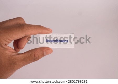 A close-up image of a hand holding a small white paper card with the word “Recruitment” in bold blue text, isolated against a clean wooden background. This minimal and professional concept photo represents hiring process, human resources, talent acquisition, job vacancy, corporate recruitment, and employment strategy. Ideal for content related to HR management, job search, career opportunities, staffing solutions, workforce planning, and business growth through talent acquisition.