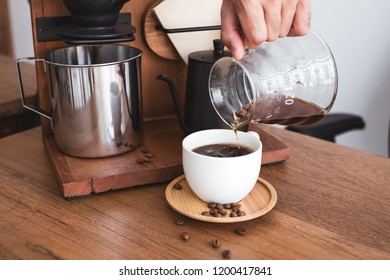 Closeup image of a hand pouring drip coffee into a white mug on vintage wooden table