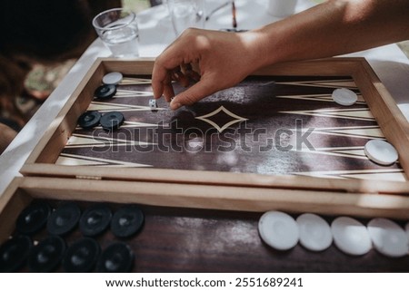 A close-up image of a hand rolling dice in a backgammon game outdoors. The scene captures the sunlight and the strategic nature of the board game.