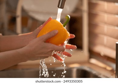 Close-up image of hands washing a yellow bell pepper under running water in a kitchen sink. The sunlight coming through the window highlights the water droplets - Powered by Shutterstock