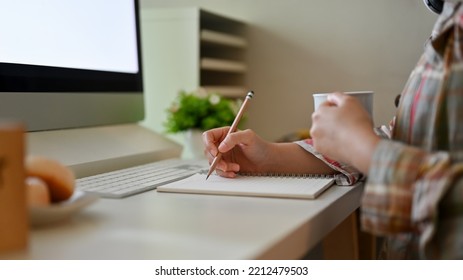 Close-up Image, A Hipster Female In A Flannel Shirt Writing Something In Her Notebook And Sipping Coffee While Sitting At Her Desk.