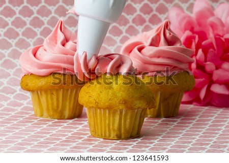 Close-up image of icing being applied to cupcakes with flower in background.