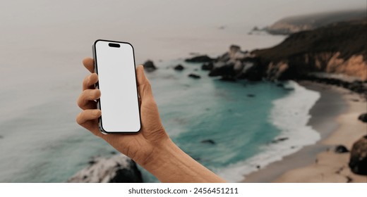 A close-up image of a male hand holding a smartphone with a blank screen, set against a blurred background featuring a serene coastal landscape with sandy beach - Powered by Shutterstock