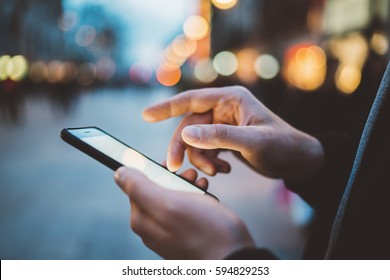 Close-up image of male hands using smartphone at night on city shopping street, searching or social networks concept, hipster man typing an sms message to his friends