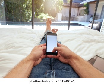 Closeup image of man lying on a bed holding a mobile phone with blank screen. POV shot of man relaxing in bedroom using touch screen cellphone.