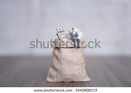 Closeup image of miniature businessman sitting on a large sack overflowing with U.S. dollar bills. Copy space for text. Success, profit, capitalist concept.