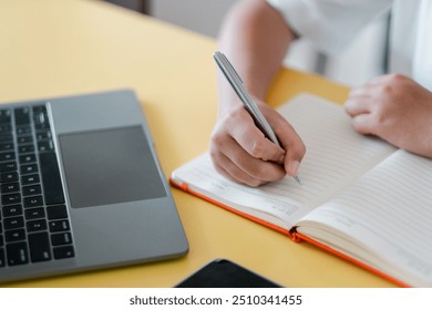 A close-up image of a person writing in a notebook with a laptop on a yellow desk, capturing a modern workspace setting. - Powered by Shutterstock