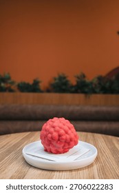Close-up image of a raspberry shaped red dessert placed on a white ceramic plate on a wooden table.