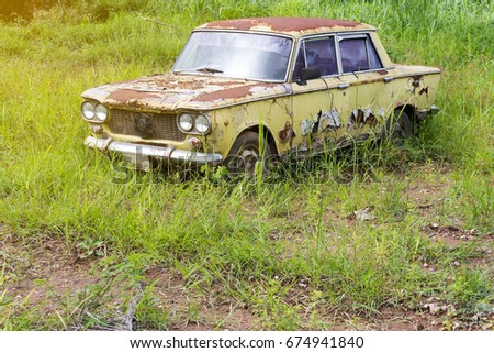 Close-up image of a rusty old green sedan parked on the ground with weed growing grass.