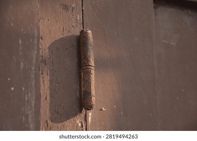 Close-up image of a rusty window hinge casting a distinct shadow on a weathered wooden window panel. The aged metal, worn paint, and warm natural sunlight emphasize the passage of time and the effects of long-term exposure to the elements. This architectural detail from an abandoned house conveys themes of decay, neglect, aging structures, restoration, and rustic heritage.