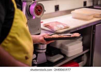 closeup image showcases the hands of a butcher carrying a tray of freshly prepared chicken nuggets, with a detailed view of a meat grinding machine in the background of his butcher shop.  - Powered by Shutterstock