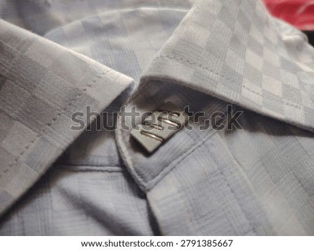A closeup image showing the inside collar of a light grey plaid shirt with a small metallic button and stitching detail