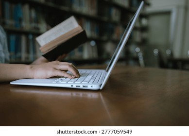 Close-up image shows a person typing on a laptop while holding a book in a library, blending digital research and reading in a quiet environment conducive to study.