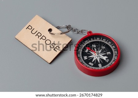 A close-up image of a small, red compass and a wooden keychain tag with the word "PURPOSE". Finding one's purpose or having a clear direction in life concept.