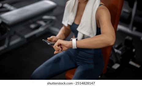 A close-up image of a sporty Asian woman in athletic wear is checking her workout results on her smartwatch while resting on a bench press at the gym. gadget and sport lifestyle concepts - Powered by Shutterstock