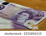 A closeup image of a stack of twenty pound banknotes featuring Queen Elizabeth II on the front, with a wooden surface in the background.