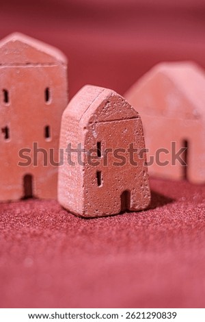 A close-up image of three small clay house models placed on a textured red surface. The shallow depth of field emphasizes the central miniature house with fine surface details. Selective focus.