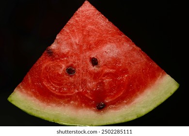 A close-up image of a triangular slice of red watermelon with visible black seeds. The juicy red flesh of the watermelon is contrasted against a dark background, highlighting its vibrant color