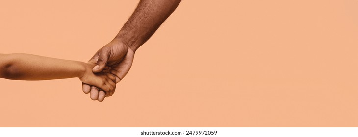 A close-up image of two hands holding each other in front of a peach-colored background. The hands are of different skin tones, representing unity and connection, panorama with copy space