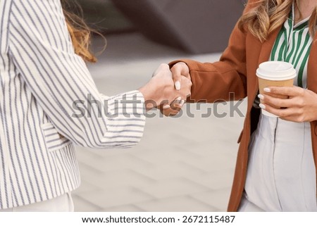 A close-up image of two professionals shaking hands in an outdoor setting formally dressed, indicating business agreement or introduction. One person holds a takeaway coffee cup, adding casual warmth
