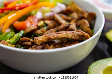 Closeup Image Of A Vegan Burrito Bowl, With Seitan As The Meat Substitute, Served Rice, Lettuce, Red And Yellow Bell Peppers, And A Sliced Avocado On The Side.